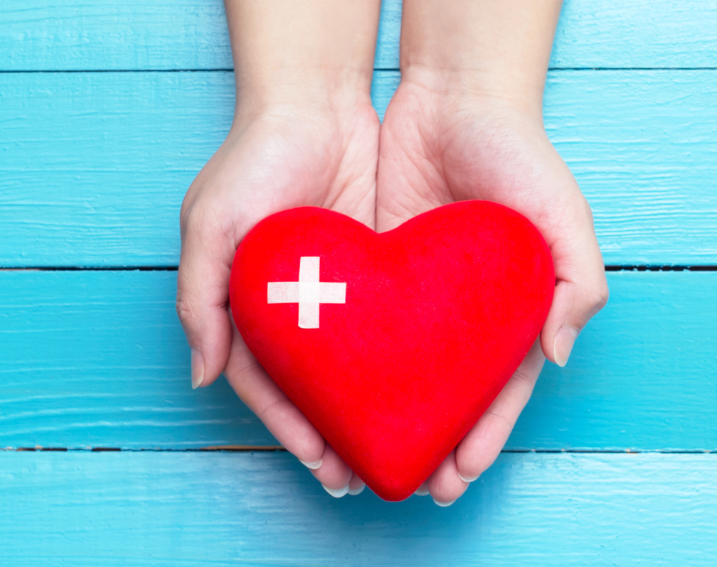 image of two hands holding a red heart with a white cross against a blue painted background