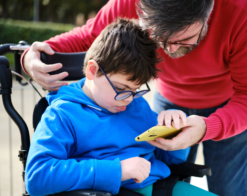 photo of a child with additional needs in a wheelchair being helped by an adult using a phone
